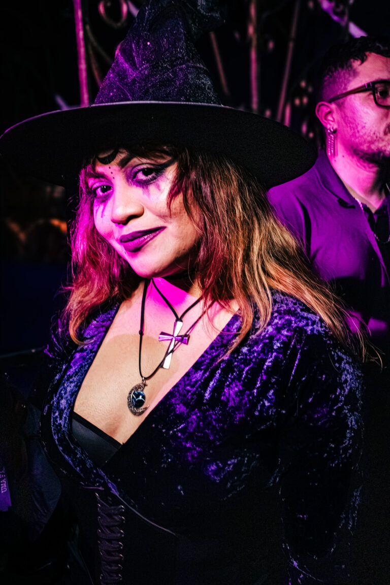 Bruxa de vestido roxo e chapéu sob luz rosa — Sad Baile Curitiba Halloween, 10/10/2026