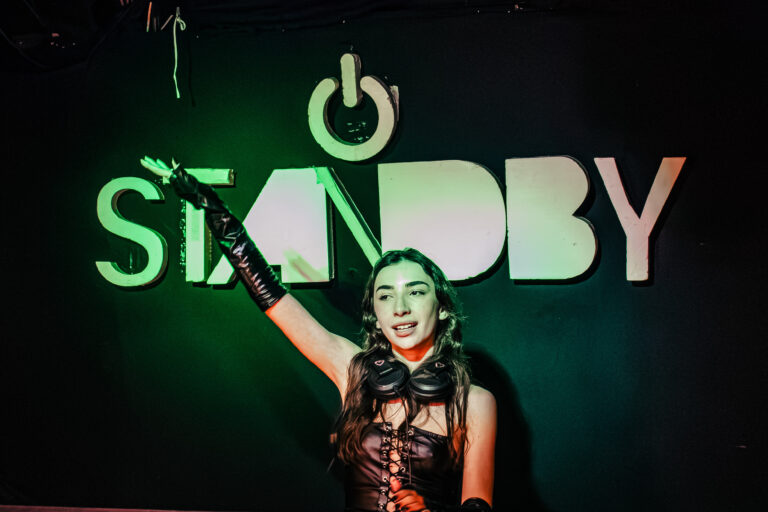 DJ levanta o braço sob o letreiro verde do Standby — Sad Baile Curitiba Halloween, 10/10/2026