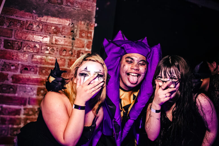 Amigas e pessoa de capuz roxo com chifres riem juntas — Sad Baile Curitiba Halloween, 10/10/2026