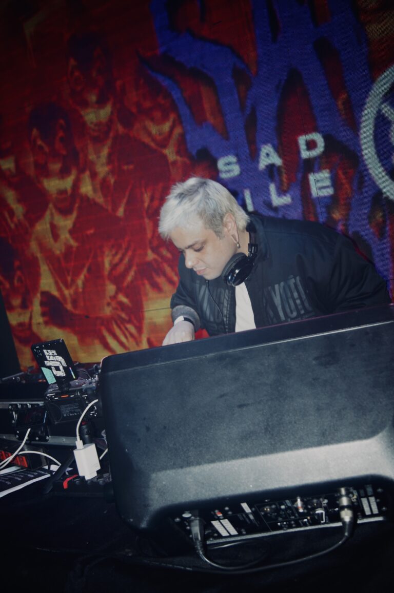 DJ de cabelo platinado toca diante do telão grafitado — Sad Baile Joker, Halloween em São Paulo, 17/10/2026