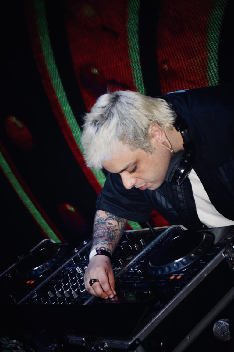 DJ de cabelo platinado inclinado sobre o mixer — Sad Baile Joker, Halloween em São Paulo, 17/10/2026