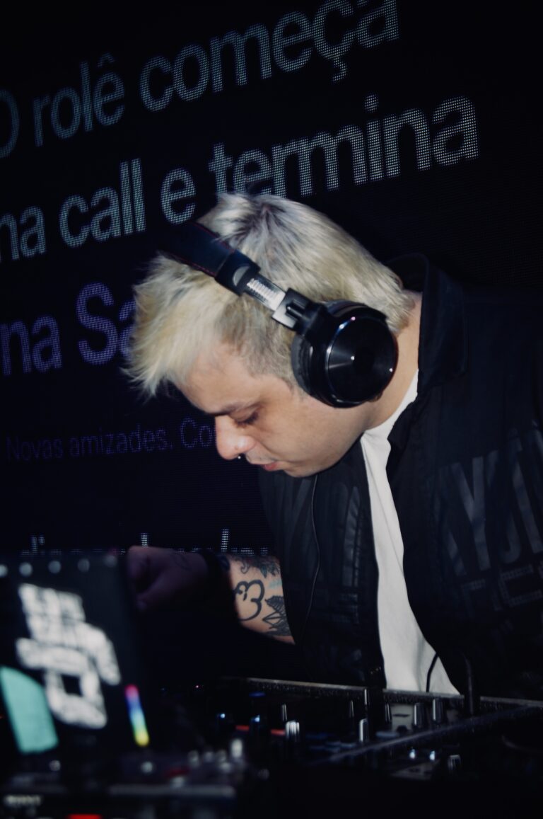 DJ de cabelo platinado diante do telão com o texto da festa — Sad Baile Joker, Halloween em São Paulo, 17/10/2026