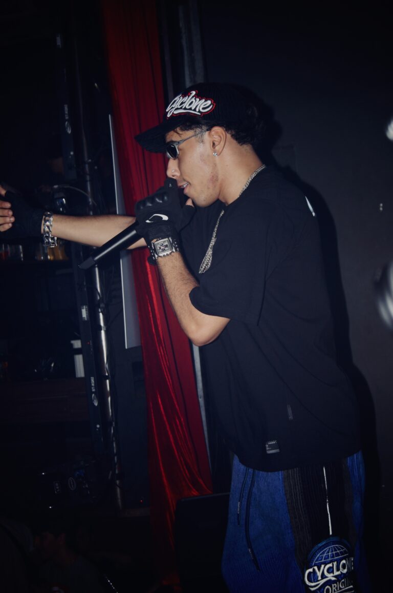 Rapper de boné canta ao microfone perto da cortina vermelha — Sad Baile Joker, Halloween em São Paulo, 17/10/2026