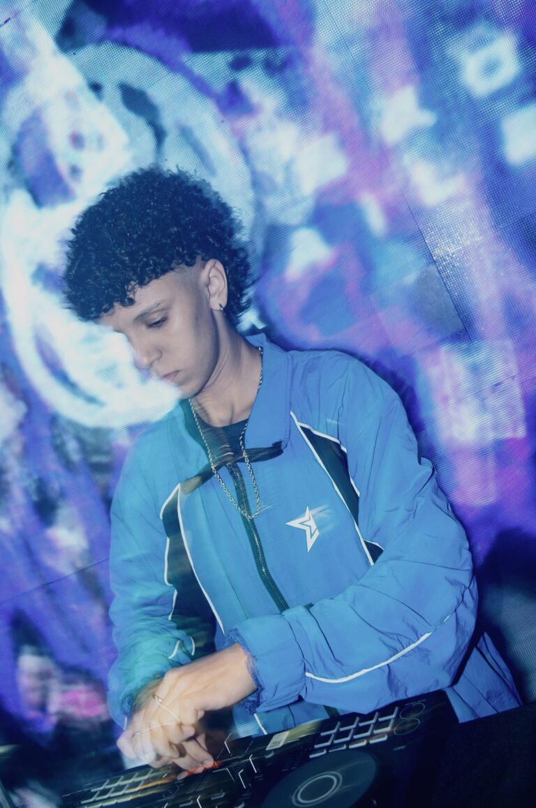 DJ de jaqueta azul diante do telão colorido, em outro ângulo — Sad Baile Joker, Halloween em São Paulo, 17/10/2026