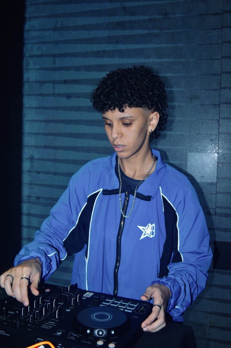 DJ de cabelo cacheado nos controles sob luz azul — Sad Baile Joker, Halloween em São Paulo, 17/10/2026