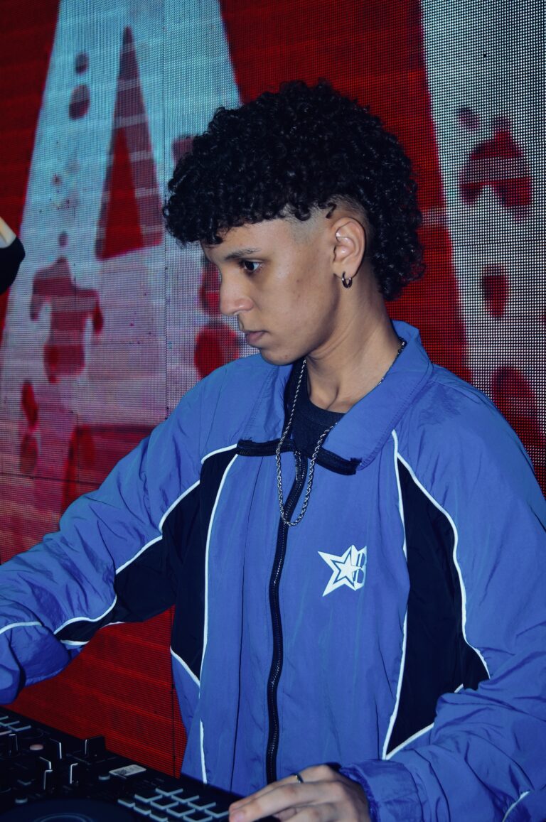 DJ de cabelo cacheado diante do telão vermelho — Sad Baile Joker, Halloween em São Paulo, 17/10/2026