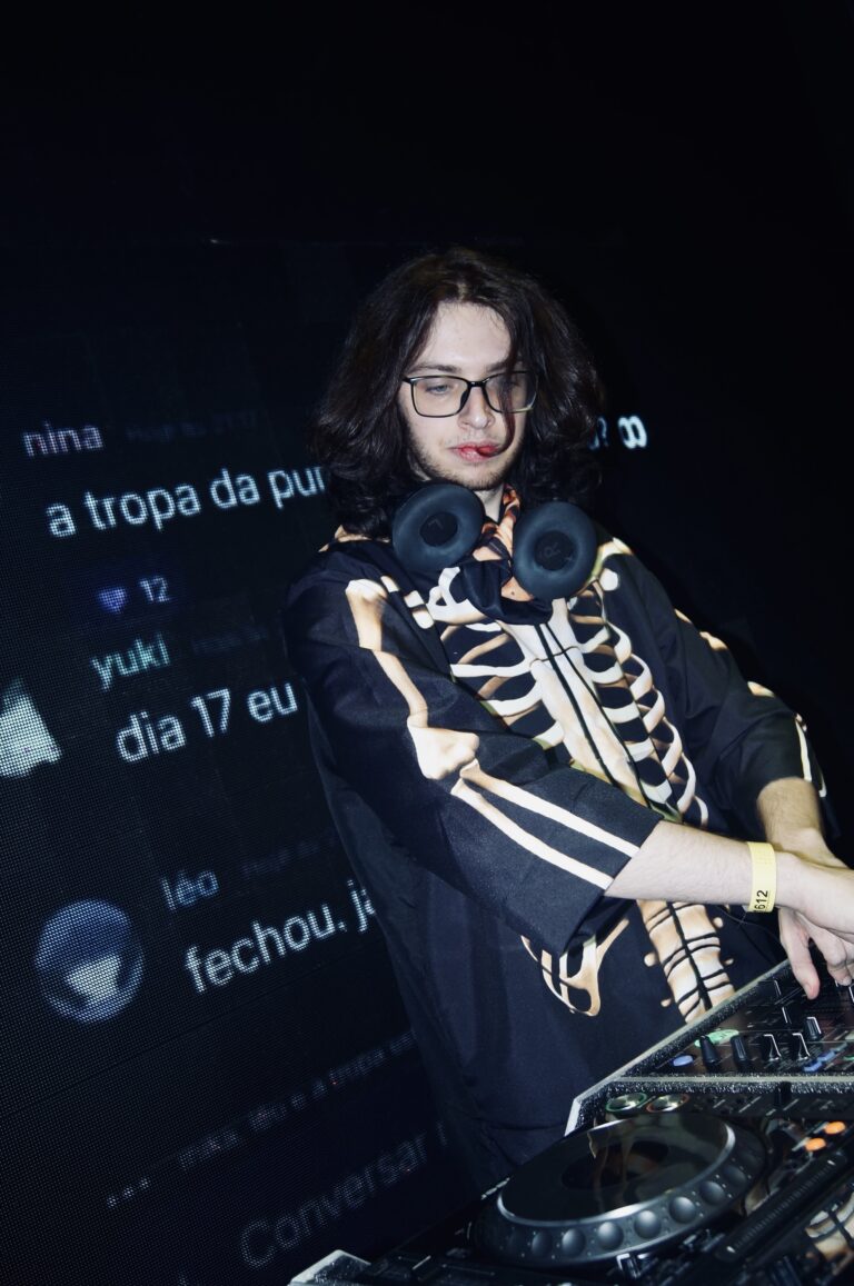 DJ de óculos e jaqueta de esqueleto diante das mensagens no telão — Sad Baile Joker, Halloween em São Paulo, 17/10/2026
