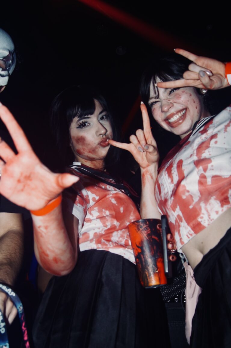 Amigas de colegial ensanguentado fazem sinal com as mãos — Sad Baile Joker, Halloween em São Paulo, 17/10/2026