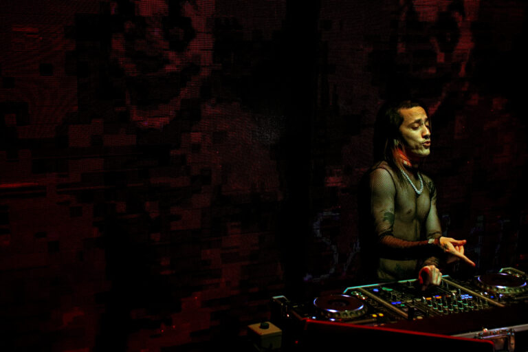 DJ tatuado na cabine com telão vermelho escuro ao fundo — Sad Baile Joker, Halloween em São Paulo, 17/10/2026