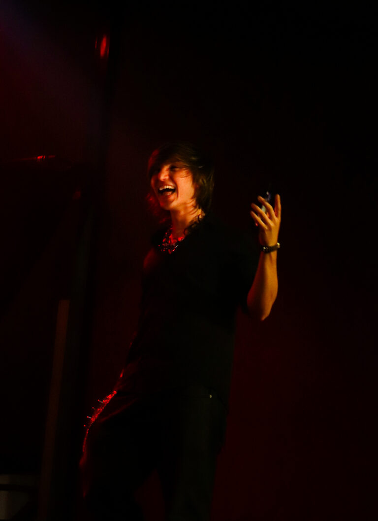 Artista de cabelo escuro canta sob luz vermelha — Sad Baile Joker, Halloween em São Paulo, 17/10/2026