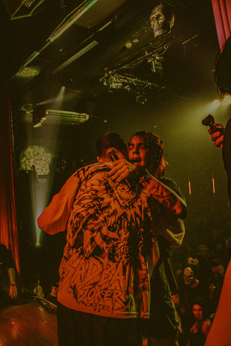 Abraço de dois artistas no palco sob luz amarela — Sad Baile Joker, Halloween em São Paulo, 17/10/2026