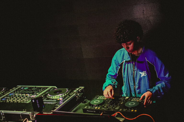 DJ de jaqueta azul mixando na penumbra — Sad Baile Joker, Halloween em São Paulo, 17/10/2026