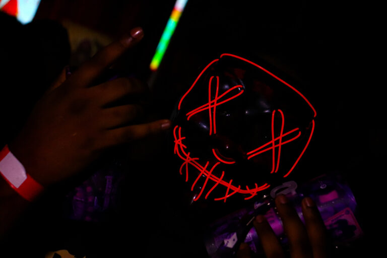 Close da máscara de néon com X e pulseira vermelha — Sad Baile Joker, Halloween em São Paulo, 17/10/2026