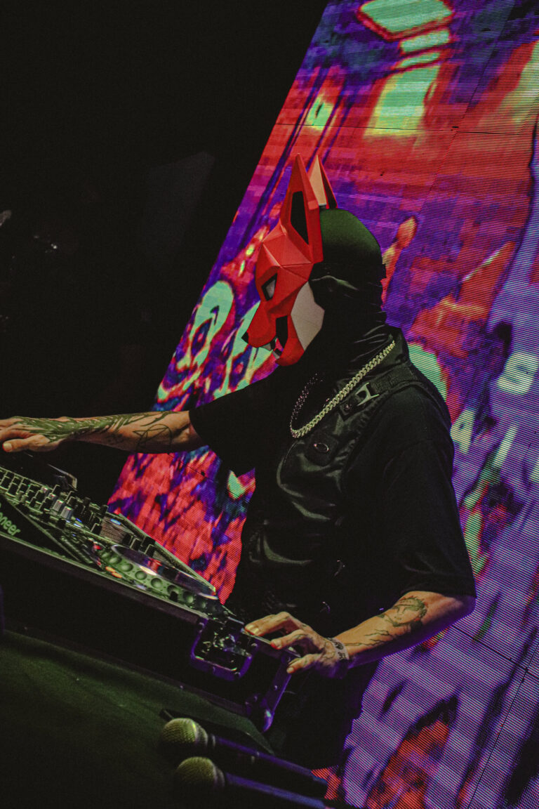 DJ mascarado inclinado sobre a mesa com telão colorido — Sad Baile Joker, Halloween em São Paulo, 17/10/2026