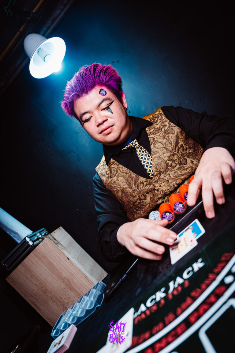 Crupiê de cabelo roxo embaralha na mesa de jogo — Sad Baile Joker, Halloween em São Paulo, 17/10/2026