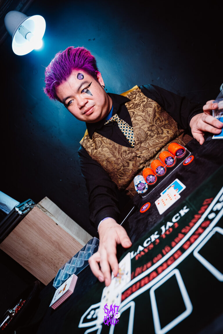 Crupiê de cabelo roxo vira as cartas — Sad Baile Joker, Halloween em São Paulo, 17/10/2026